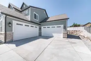Exterior of a house with concrete driveway and two closed white garage doors with windows. Side view of a gray large house with bricks and rock landscape and fence on the side.