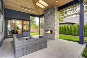 New modern home features a backyard with covered patio accented with stone fireplace, vaulted ceiling with skylights and furnished with gray wicker sofa placed on concrete floor.